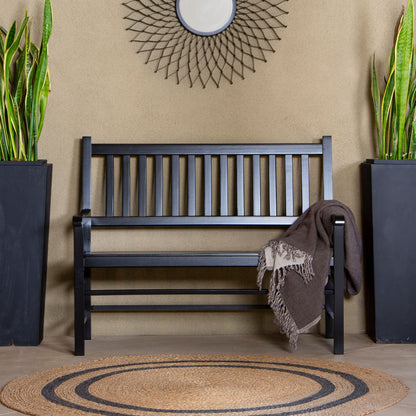 A black wooden slat bench is centered against a beige wall, with a brown throw blanket draped over one arm. Two tall black planters with green snake plants flank the bench, and a round woven rug sits in front. 