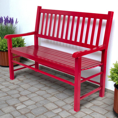 A bright red wooden garden bench with a slatted back and seat, shown outdoors on a stone patio against a white wall with potted plants nearby. 