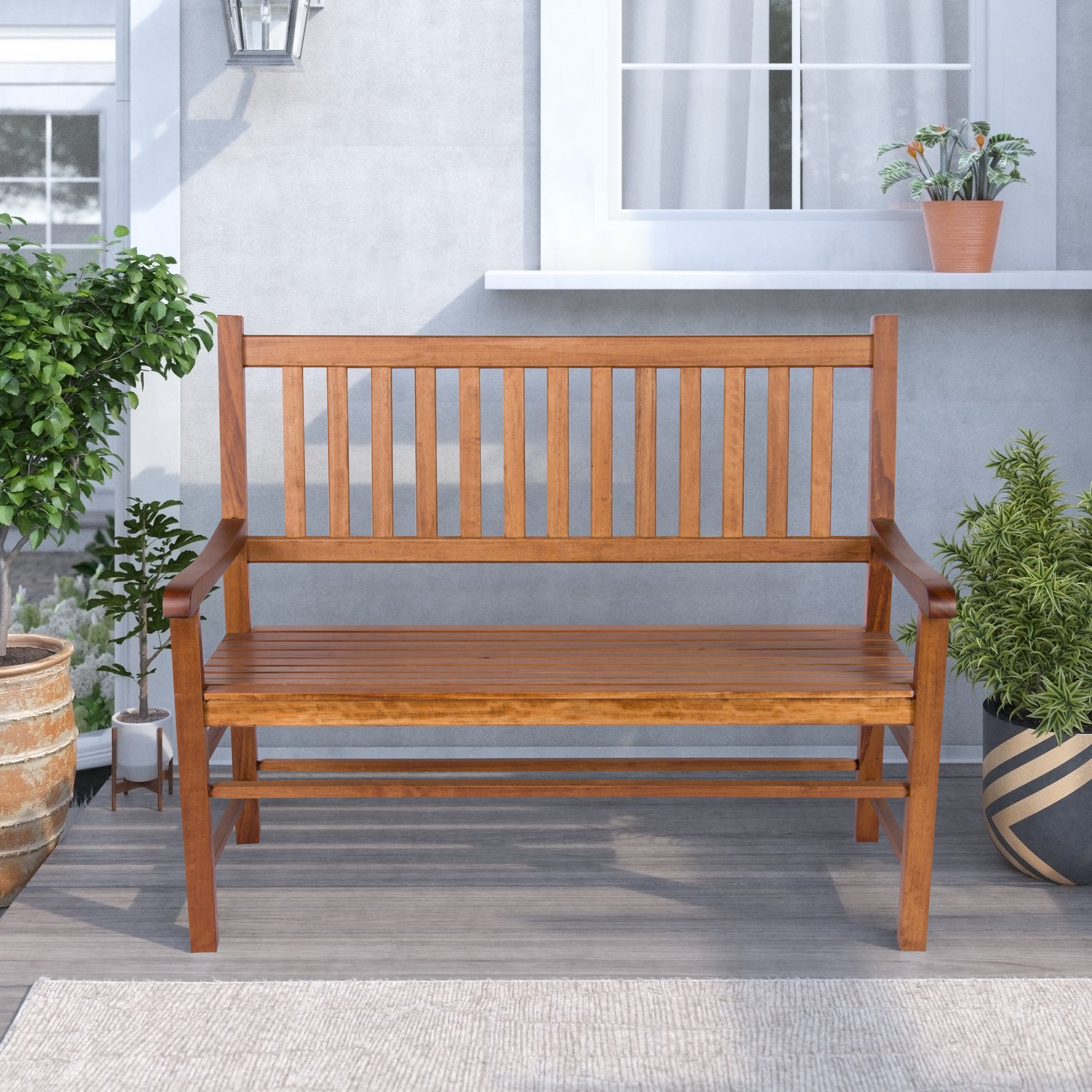 A wooden outdoor bench with a slatted back and seat, shown in a patio setting with potted plants and a gray stucco wall background. 