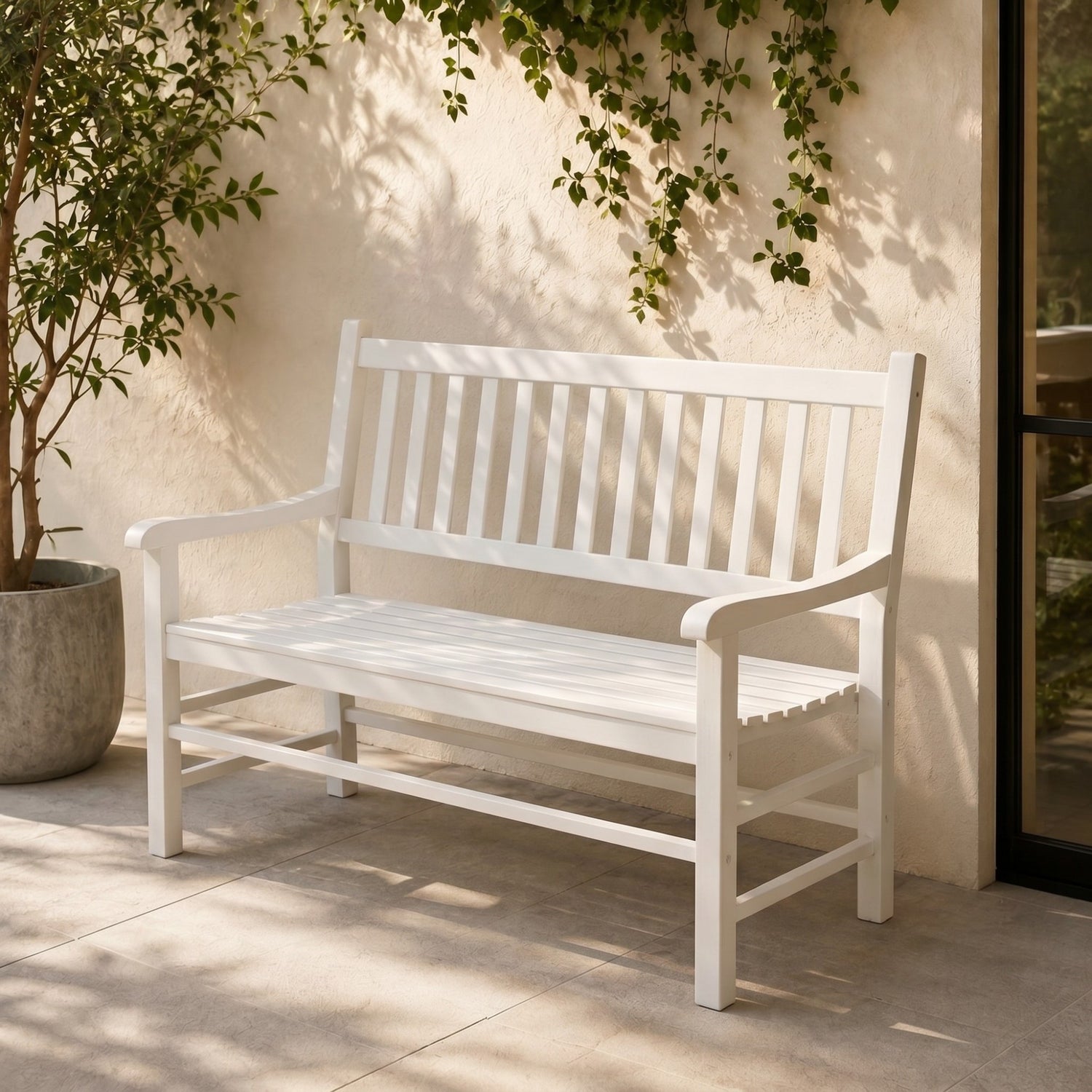 A white wooden outdoor bench with a slatted back and seat, placed on a light stone patio against a beige stucco wall with climbing green vines. 