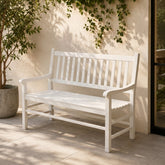 A white wooden outdoor bench with a slatted back and seat, placed on a light stone patio against a beige stucco wall with climbing green vines. 