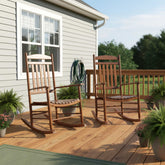 Two wooden rocking chairs on a wooden deck in front of a house with light gray siding, with potted plants and a garden hose in the background. 