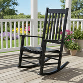 A black rocking chair on a wooden deck with a white railing and garden plants in the background. 