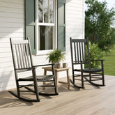 Two black rocking chairs on a wooden front porch beside a small side table with a potted plant and mug, set against a white house with green shutters. 