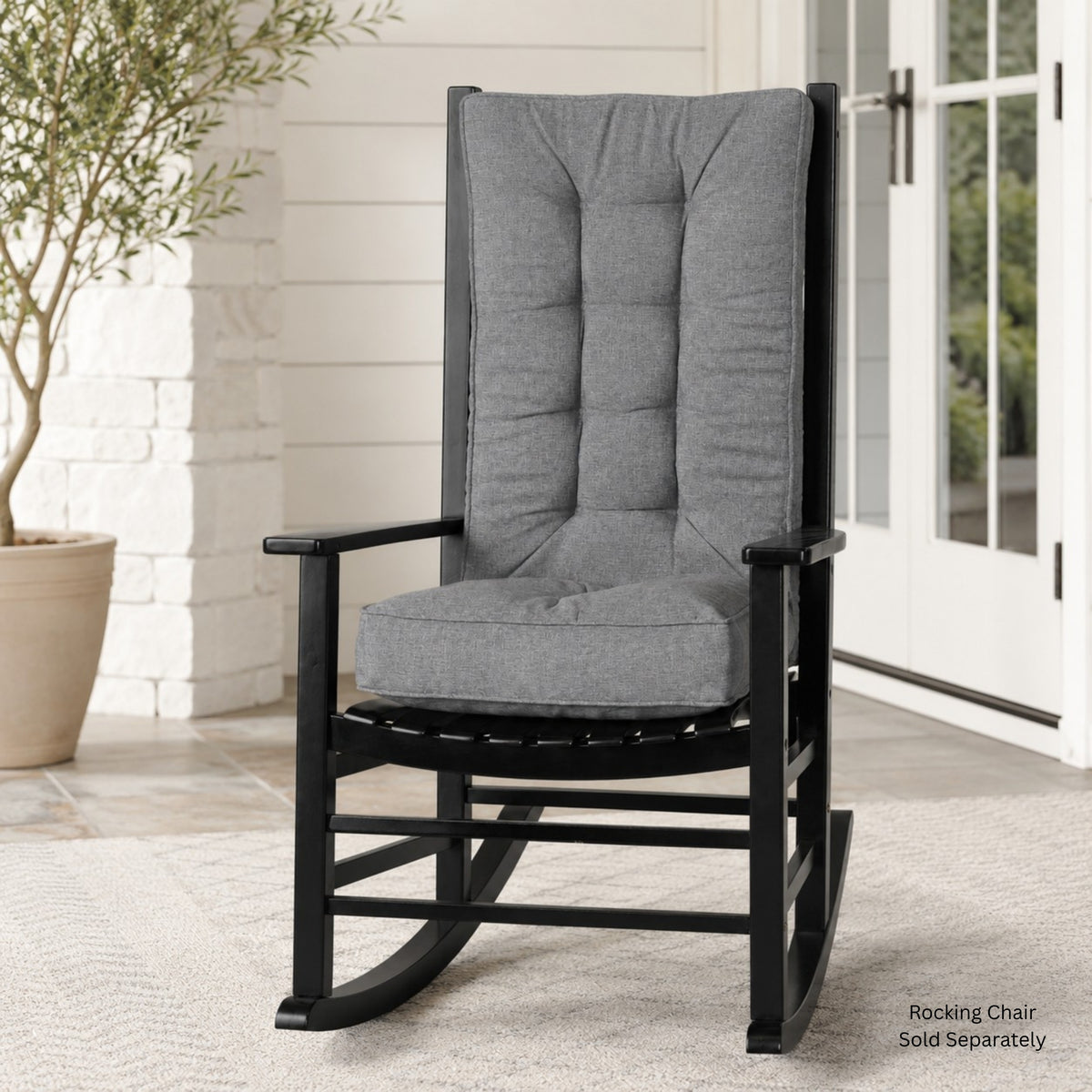 A black rocking chair with a tall, high-back tufted gray cushioned backrest and seat cushion, shown on a patio porch next to a potted tree. Text in the bottom right corner reads &