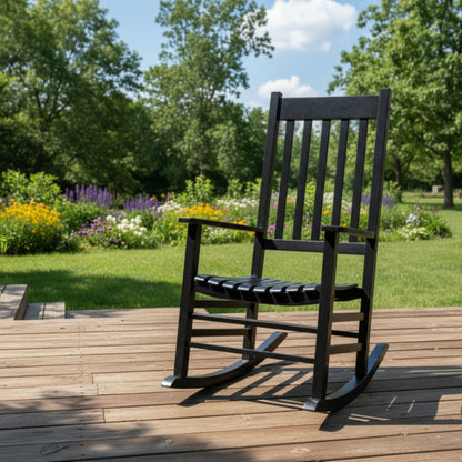 A black outdoor rocking chair with a slatted back and seat, placed on a wooden deck with a lush green garden and trees in the background. 
