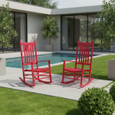 Two bright red rocking chairs placed on a concrete patio beside a swimming pool in a modern backyard setting with green grass and shrubs. 