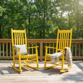 Two bright yellow outdoor rocking chairs on a wooden deck with a green forest background. One chair has a striped pillow, and the other has a beige throw blanket. 