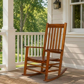 A wooden rocking chair with a warm brown finish sits on a porch next to a white column, with a green lawn and trees visible in the background. 