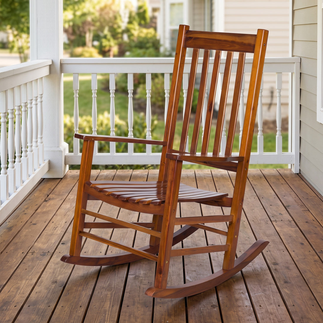 A wooden rocking chair with a warm brown finish sits on a wooden porch floor with white railings and a blurred outdoor background. 