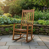 A classic wooden rocking chair with a slatted back and seat, positioned on a stone patio in a lush, green garden setting. 
