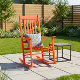 An outdoor patio scene with a bright orange rocking chair as the main product, styled with a blue and white patterned throw pillow. A small dark side table sits beside it on a light gray tiled patio, with a green lawn, plants, and a wooden fence in the background. 