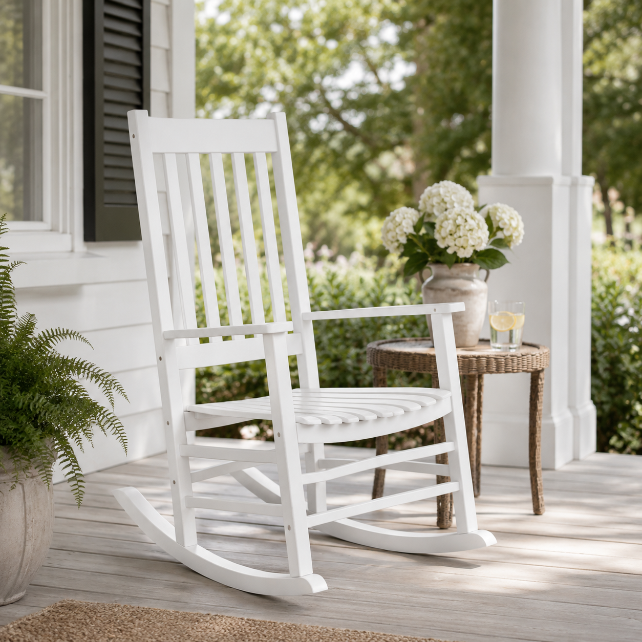 A white wooden rocking chair sits on a porch next to a small round side table with white flowers, with a potted fern in the foreground and a white house exterior behind it. 