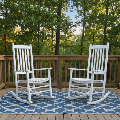 Two white wooden rocking chairs placed on a blue and white patterned outdoor rug on a wooden deck, with a forest background visible behind a  railing. 