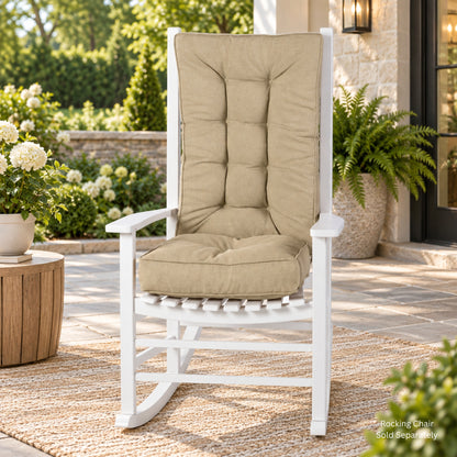 A white wooden rocking chair with thick, tufted beige cushions sits on a woven rug in a beautifully landscaped outdoor patio setting with potted plants and a stone wall. 