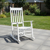 A white outdoor rocking chair with a slatted back and seat, placed on a light gray tiled patio in a backyard with green grass and a wooden fence in the background. 