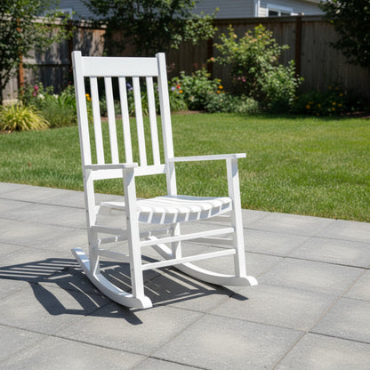 A white outdoor rocking chair with a slatted back and seat, placed on a light gray tiled patio in a backyard with green grass and a wooden fence in the background. 