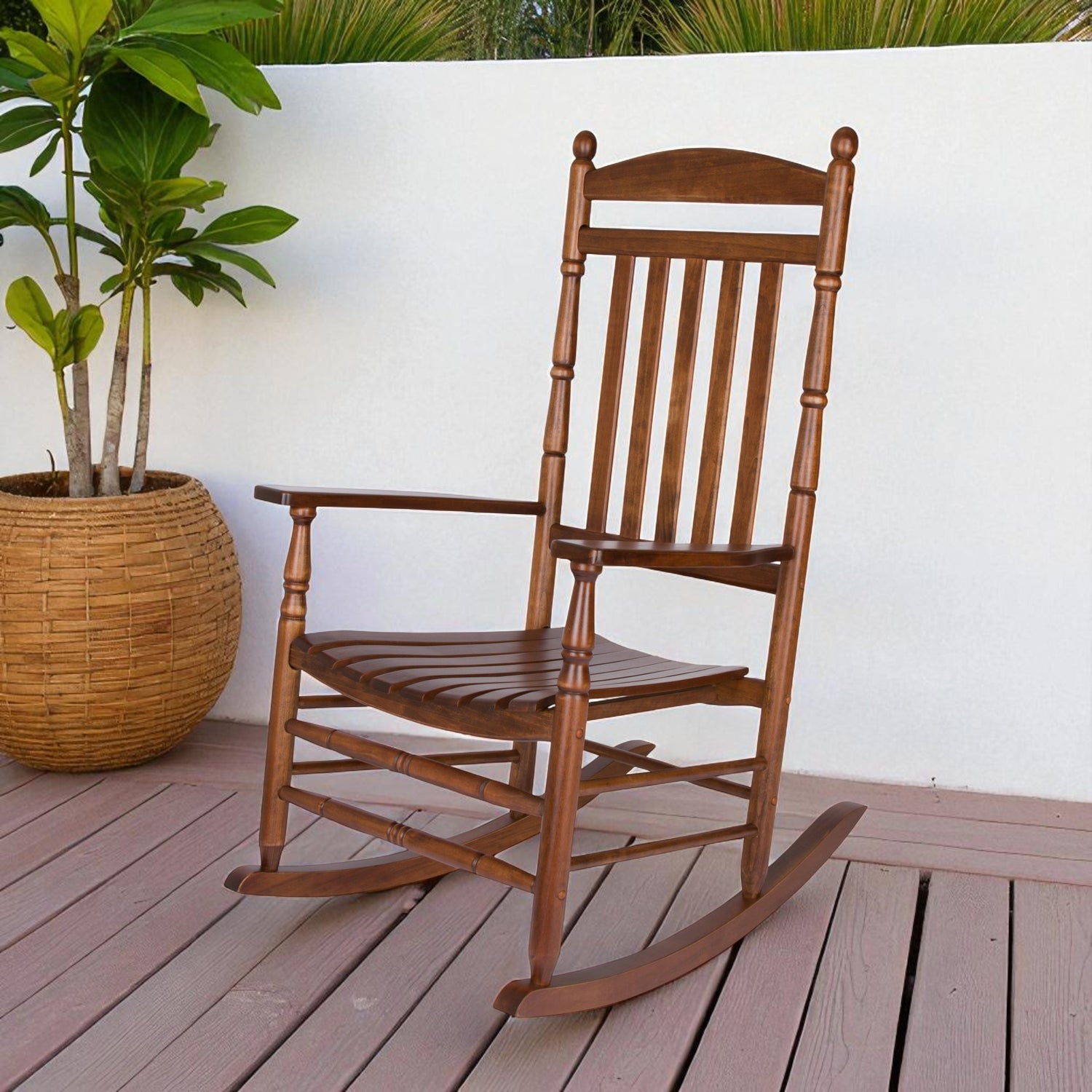 A wooden rocking chair with a warm medium-brown finish, set on a wooden deck in front of a white wall next to a potted plant. 
