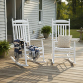 Two white outdoor rocking chairs on a wooden deck in front of a light gray house. One chair has a navy, beige, and white striped throw blanket draped over it, and the other has a beige chevron-patterned pillow. 