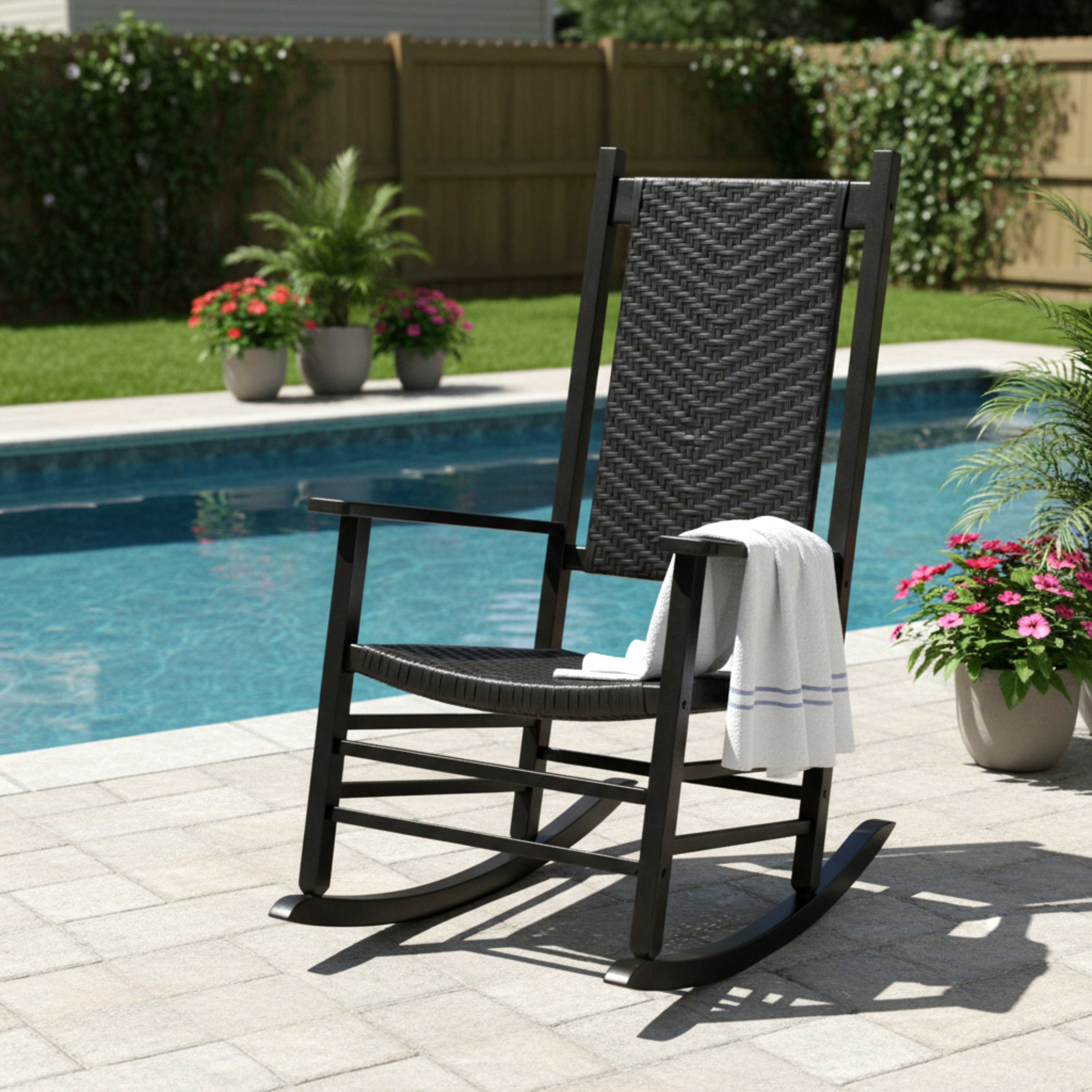 A black outdoor rocking chair with woven wicker-style seat and backrest, placed on a poolside patio with a white towel draped over the arm. A swimming pool, grass, fence, and potted plants are visible in the background. 