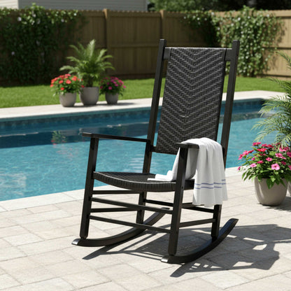 A black outdoor rocking chair with woven wicker-style seat and backrest, placed on a poolside patio with a white towel draped over the arm. A swimming pool, grass, fence, and potted plants are visible in the background. 