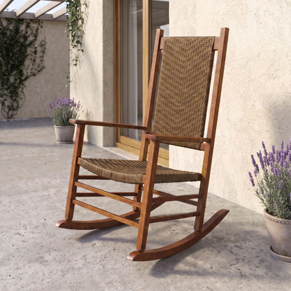 A wooden rocking chair with woven wicker-style seat and back slats, shown outdoors on a patio beside a stucco wall, glass door, and potted lavender plants. 