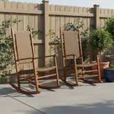 Two wooden rocking chairs with woven rope seats and backs are placed on a concrete patio in front of a wooden fence with climbing plants and potted shrubs. 