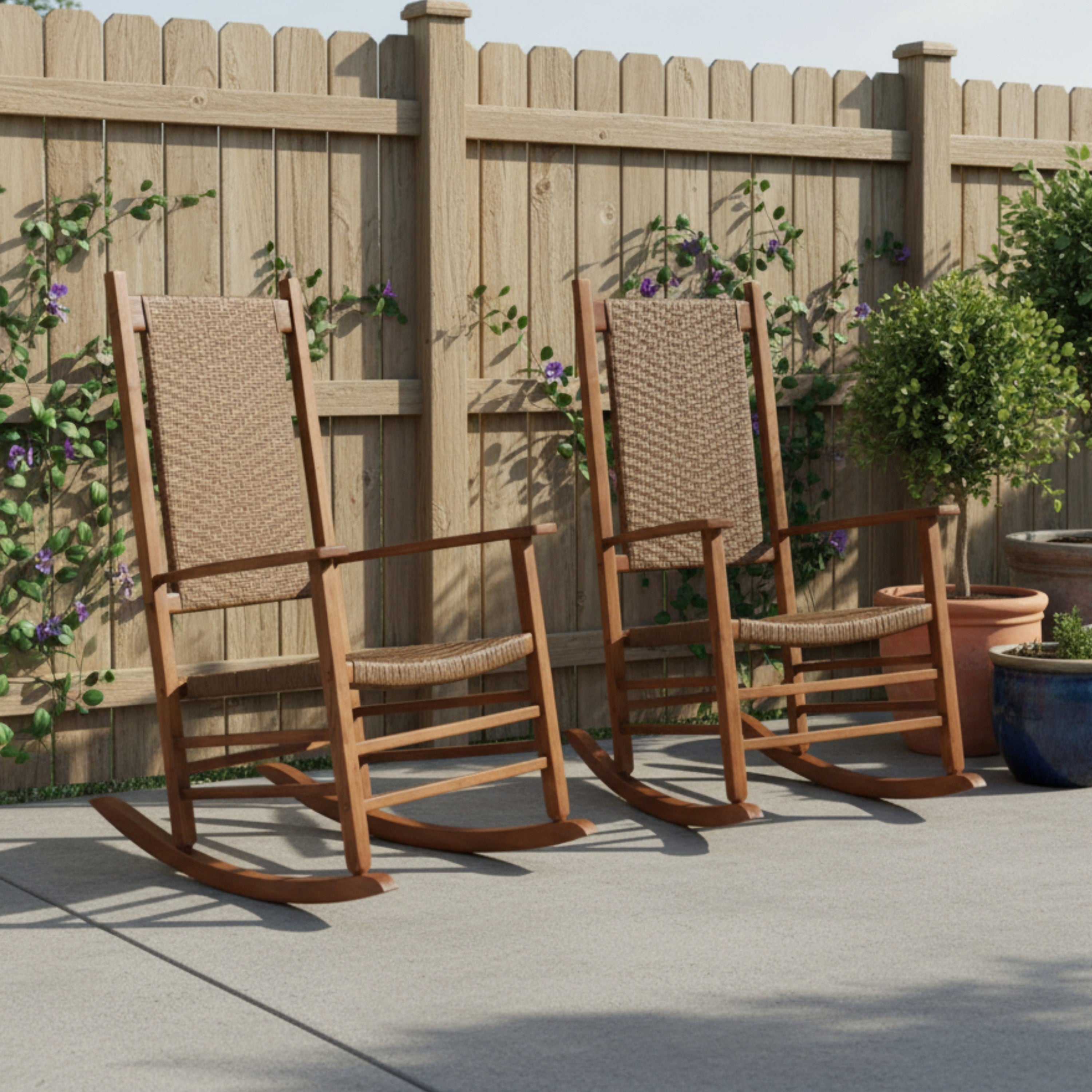 Two wooden rocking chairs with woven rope seats and backs are placed on a concrete patio in front of a wooden fence with climbing plants and potted shrubs. 