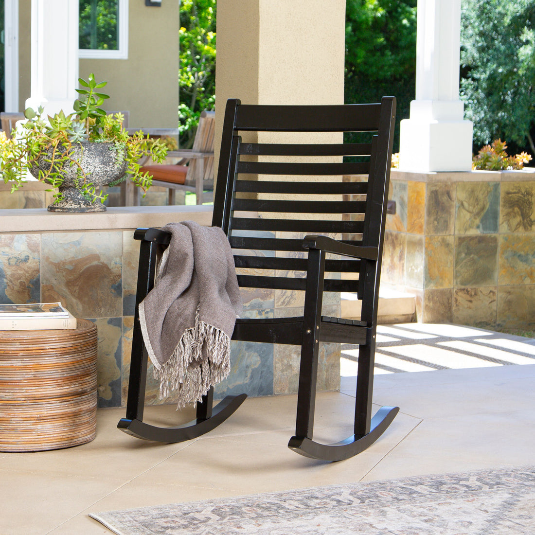 A black wooden rocking chair on a tiled porch with a gray throw blanket draped over the arm, set against a background of columns and greenery. 