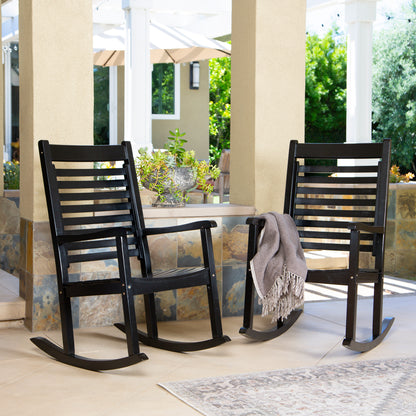 Two black outdoor rocking chairs on a covered patio with a beige throw blanket draped over one chair, columns, plants, and greenery in the background. 