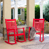 Two bright red outdoor rocking chairs on a covered patio, with a gray throw blanket draped over one chair. The setting includes beige pillars, stone walls, greenery, and a patio umbrella in the background. 