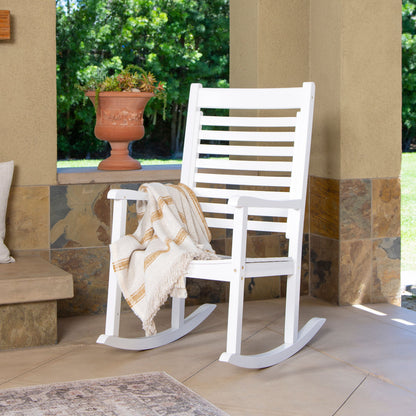 A white outdoor rocking chair with a slatted back and curved rockers, shown on a patio with a cream throw blanket draped over the seat. A terracotta planter and greenery are visible in the background. 
