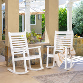 Two white outdoor rocking chairs on a covered patio, with a beige throw blanket draped over one chair. The setting includes stone columns, a tiled wall, greenery, and a patio umbrella in the background. 