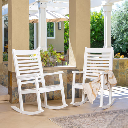 Two white outdoor rocking chairs on a covered patio, with a beige throw blanket draped over one chair. The setting includes stone columns, a tiled wall, greenery, and a patio umbrella in the background. 