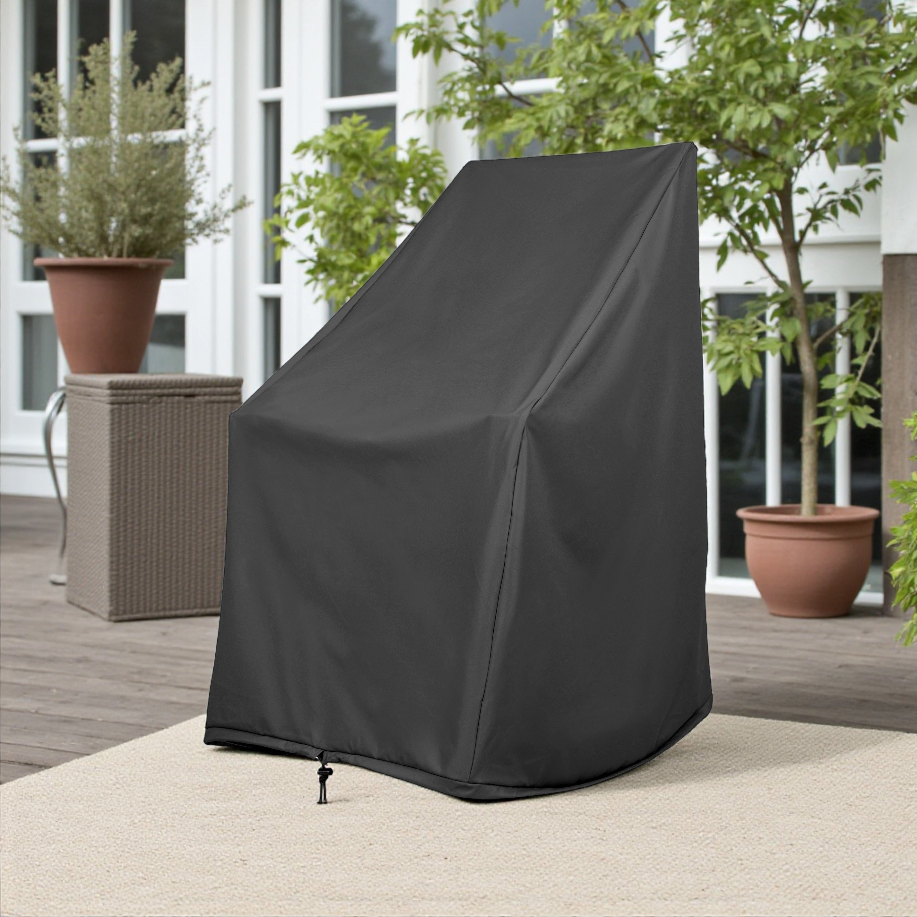 A black outdoor patio chair cover standing upright on a light beige rug on a wooden deck, with potted plants and a patio setting in the background. 