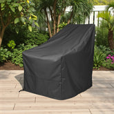 A black outdoor patio chair cover standing on a light wood deck in a garden setting with plants and trees in the background. 
