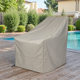 A gray patio furniture cover, likely for a lounge chair or small table, shown outdoors on a wooden deck next to a swimming pool with green shrubs and houses in the background. 