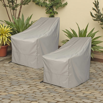 Two outdoor patio armchairs covered with light gray protective covers, placed on a stone patio in front of a beige wall with green plants. 
