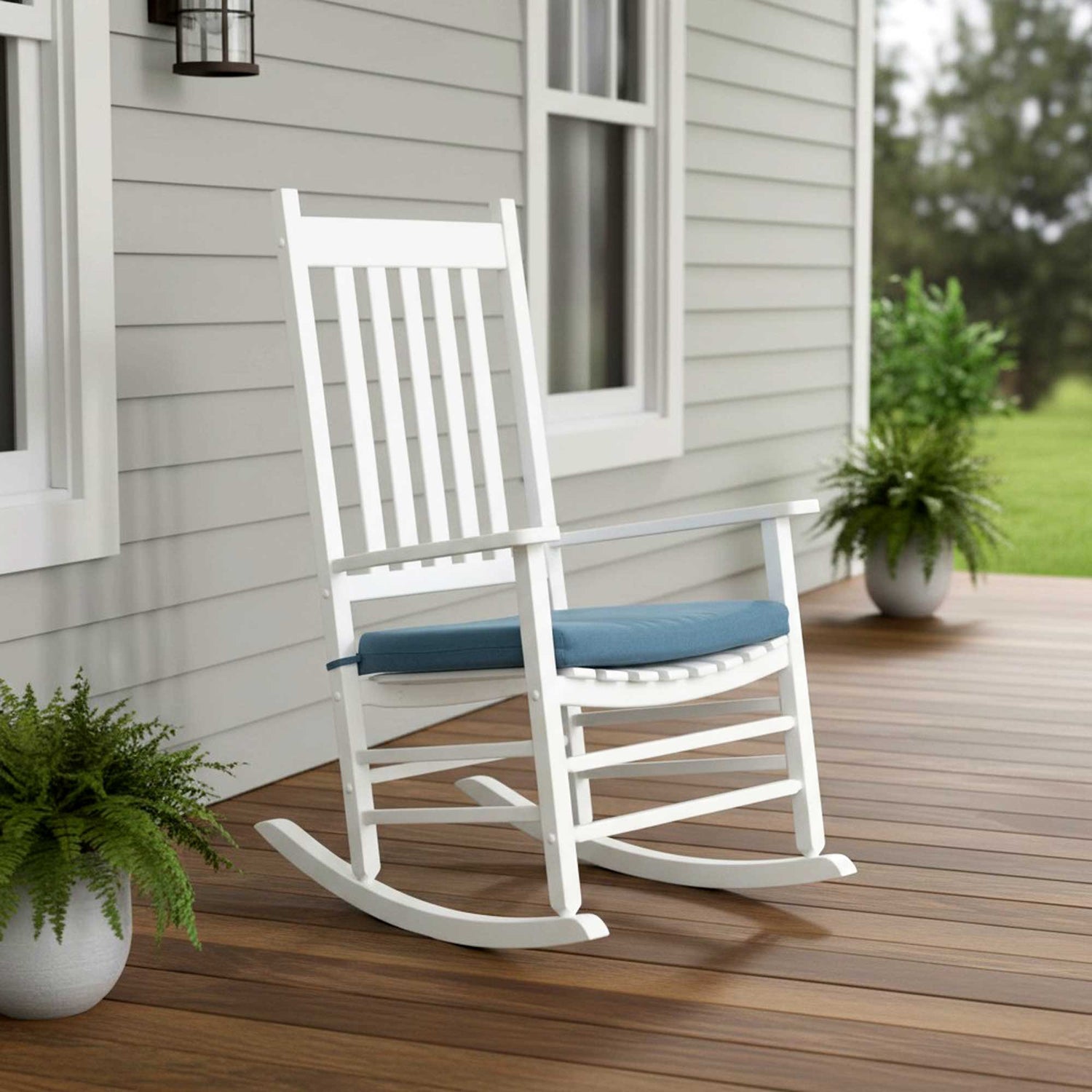 A white wooden rocking chair with a blue cushion sits on a wooden deck in front of a house with light gray siding. 
