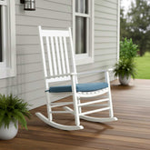 A white wooden rocking chair with a blue cushion sits on a wooden deck in front of a house with light gray siding. 