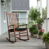 A wooden rocking chair with a black cushion sits on a gray porch deck in front of a light blue house, surrounded by potted plants. 