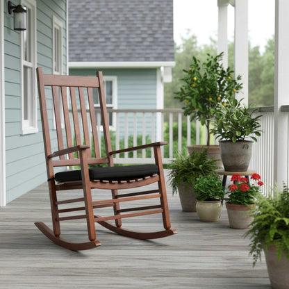 A wooden rocking chair with a black cushion sits on a gray porch deck in front of a light blue house, surrounded by potted plants. 