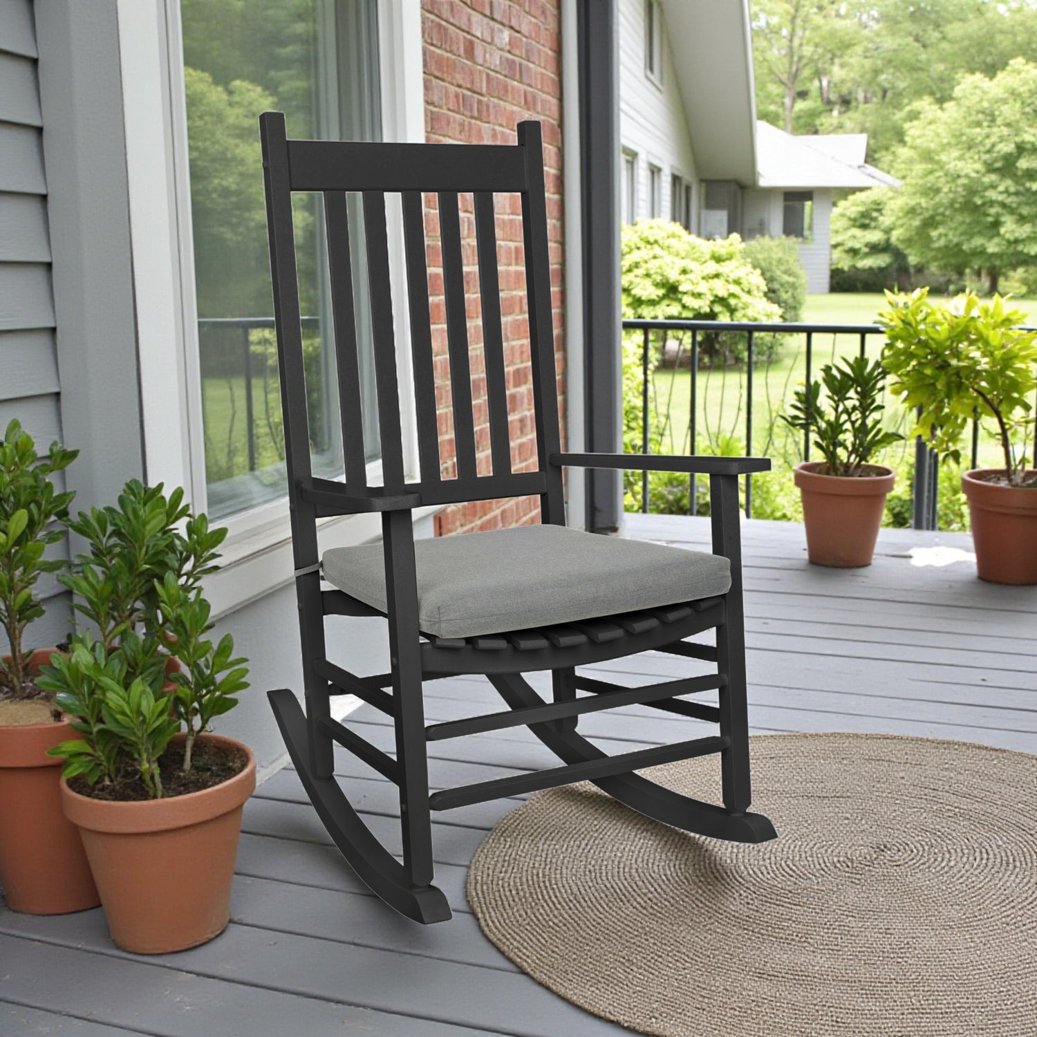 A black rocking chair with a light gray cushion sits on a gray porch next to potted green plants and a woven rug, with a brick wall and house exterior in the background. 