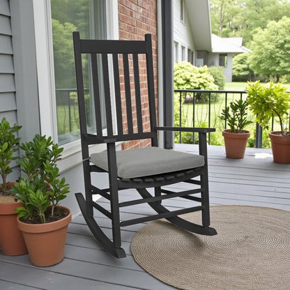 A black rocking chair with a light gray cushion sits on a gray porch next to potted green plants and a woven rug, with a brick wall and house exterior in the background. 