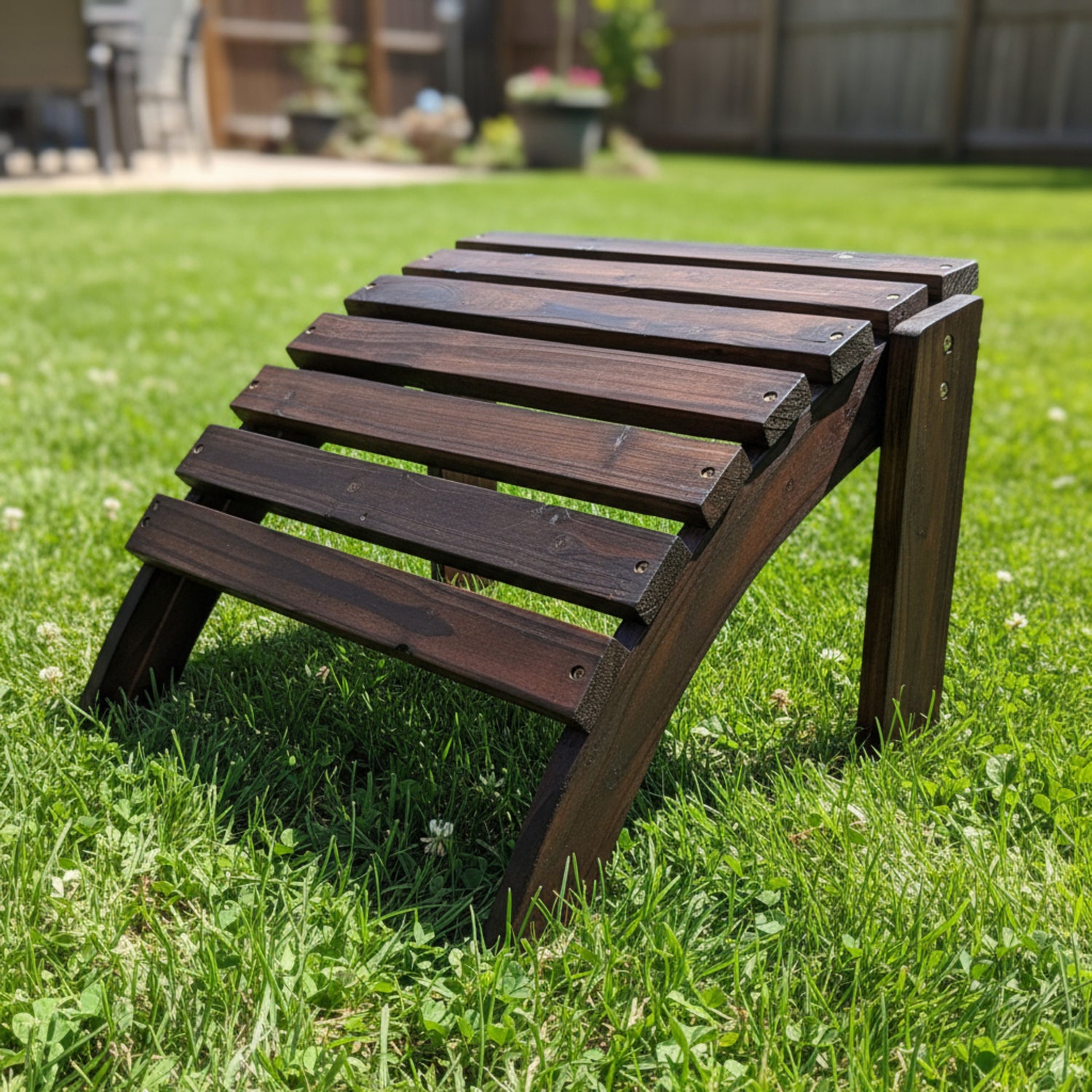 A dark brown wooden outdoor lounge chair with a slatted backrest, positioned on a green grass lawn with a blurred backyard background. 