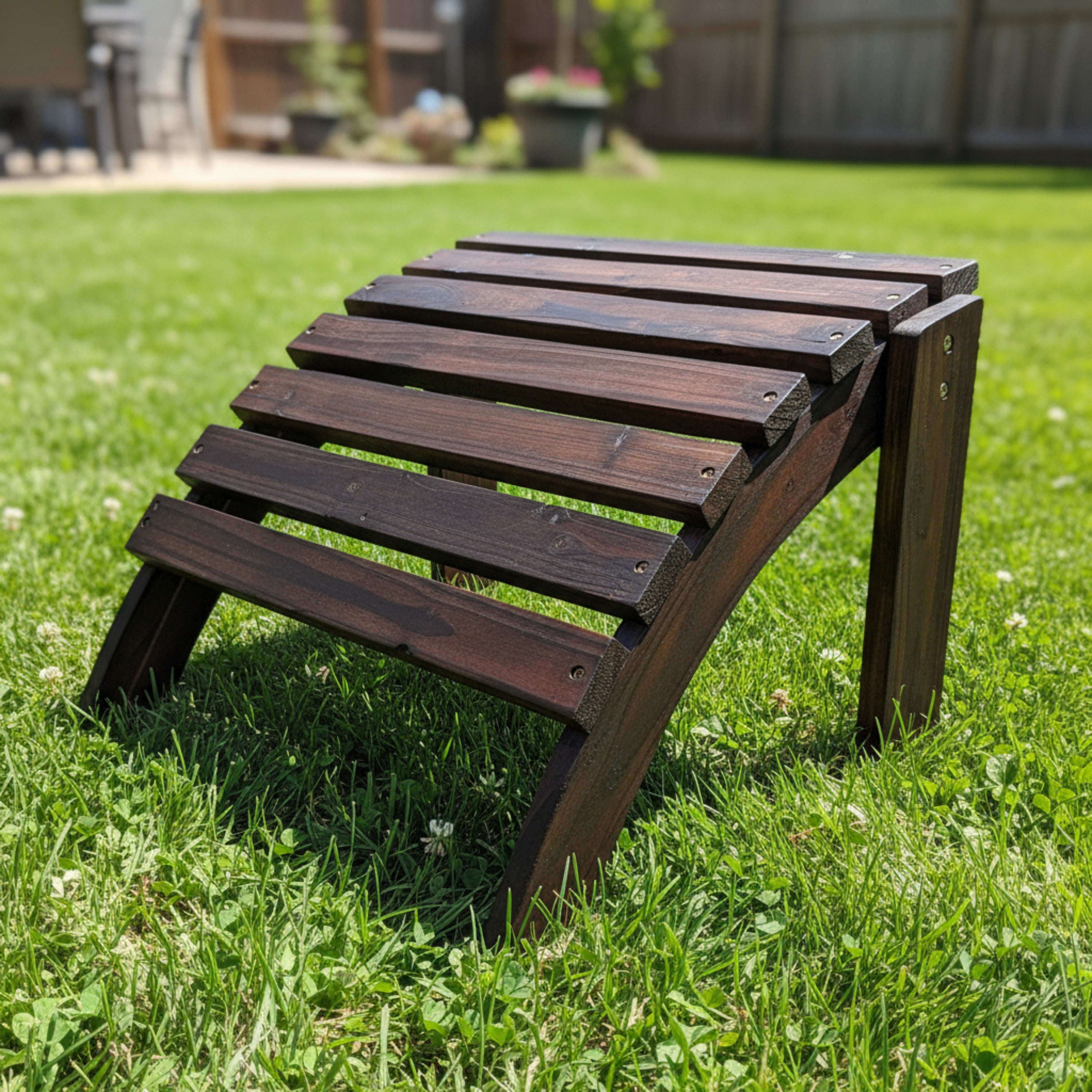 A dark brown wooden outdoor lounge chair with a slatted backrest, positioned on a green grass lawn with a blurred backyard background. 