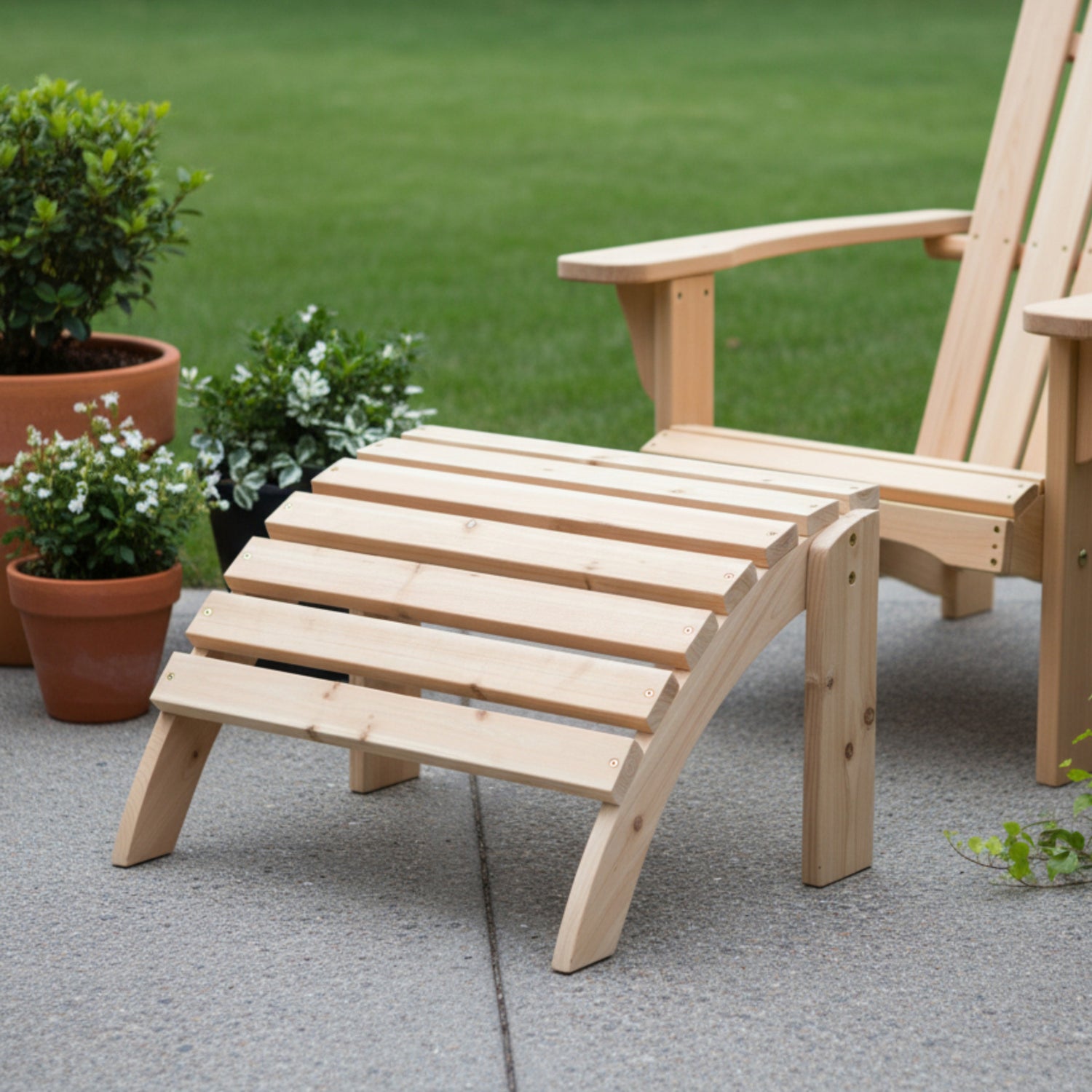A light natural wood outdoor lounge chair and matching footrest set on a concrete patio with a green lawn and potted plants in the background. 