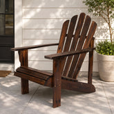 A dark brown wooden Adirondack chair placed on a light gray concrete patio in front of a white house exterior, with a potted plant to the right. 