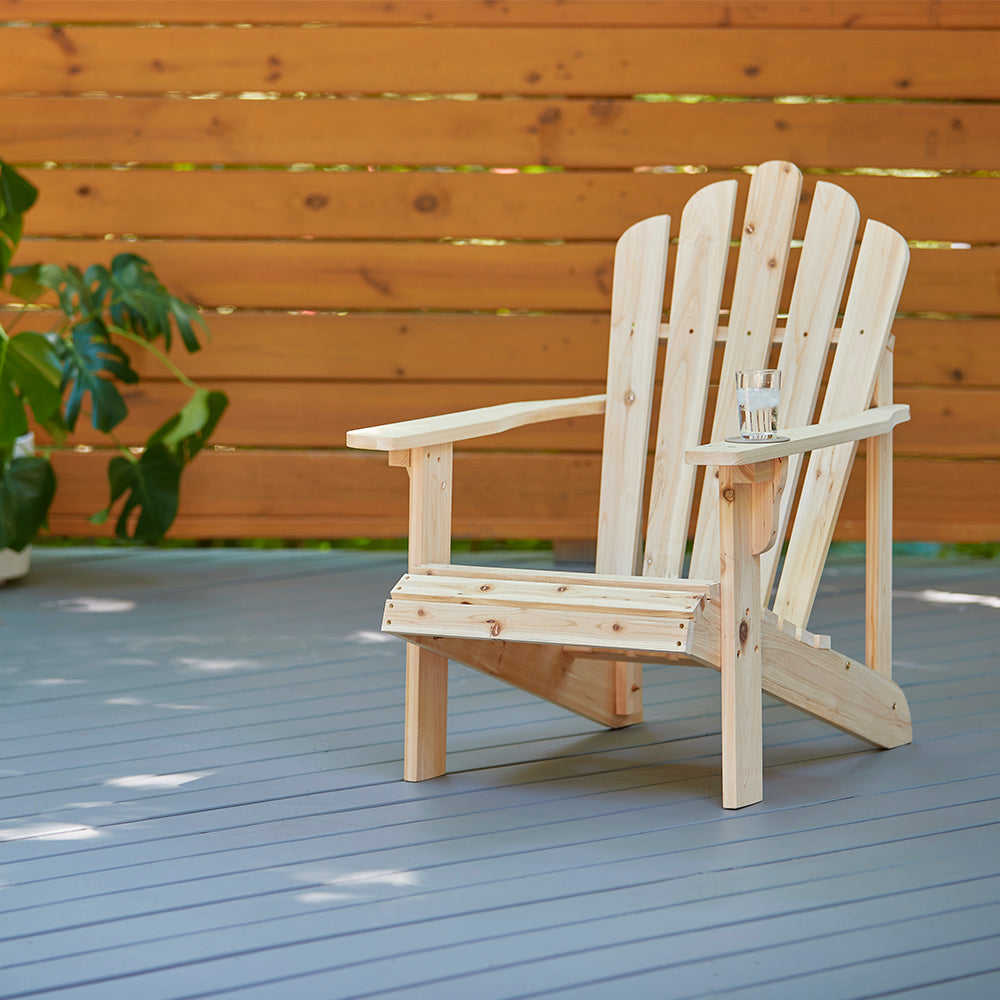 A light natural wood Adirondack chair with a small glass cup on its armrest, set on a gray wooden deck in front of a warm brown wooden fence. 
