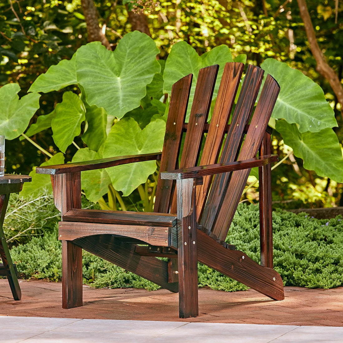 A dark brown wooden Adirondack chair placed on a patio with large green leaves and garden plants in the background. 